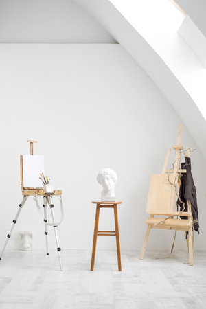 White and bright studio with a window and gray floor. Workspace of the artist. Easel, canvases and plaster figures for learning to draw. Conceptual interior without people.の写真素材