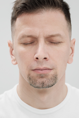 The face of a bearded brunet man in white clothes, eyes closed close up. Smooth skin, beautiful features.の写真素材