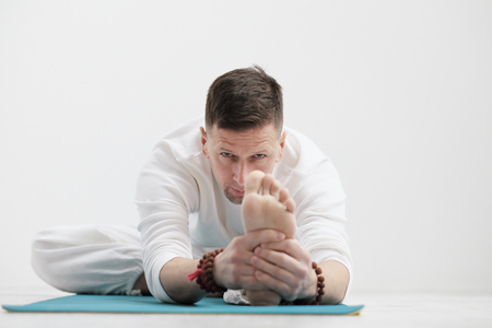 Handsome young man in white clothes does exercises. Yoga asanas and poses for stretching and meditating on a blue rug.の写真素材