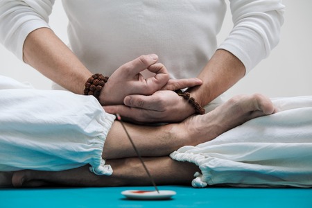 The concept of yoga and meditation. Close-up of the hands and feet of a man in white clothes on a light background. Against the background of the statue of Buddha.の写真素材