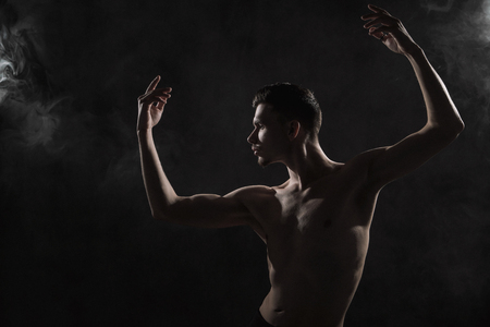 A young male ballet dancer with black leggings and a naked torso performs dance moves against a gray grunge background, with a light of lights and smoke. Conceptual art of contemporary.の写真素材