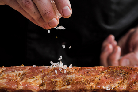 Raw piece of meat, beef ribs. The hand of a male chef puts salt and spices on a dark background, close-up.の写真素材