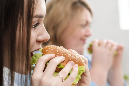 Two girls, slim and fat, blonde and brunette, eat hamburgers. On a white background, the theme of diet and proper nutrition, choice and opposition.の写真素材