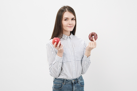 A slender beautiful brunette girl on a white background holding a pink glazed donut and a red apple in her hands.の写真素材