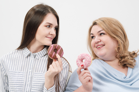 Two girls, thin and fat on a white background, are holding pink glazed donuts in their hands, smiling and joking.の写真素材