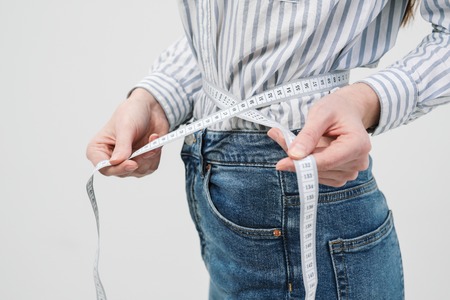 Young slim slim girl measures the waist tape in jeans and shirt on a white background. Concept of...