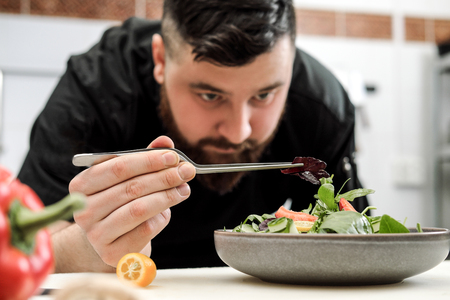 A male chef decorates a salad dish in a restaurant kitchen in a black apron. White cutting board, closeup of a hand with tongs.の写真素材