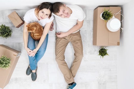 A young married couple with a red cat, a man and a woman, are sitting on the floor in a bright room against the background of cardboard boxes, green plants and things. Concept themes moving to a new home.の写真素材