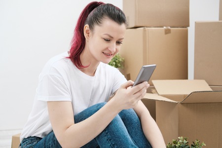 Young beautiful girl with colored hair in a white T-shirt and jeans, talking on the phone and writes messages against the background of cardboard boxes and things. The concept of transporting things on the day of moving to a new home.の写真素材