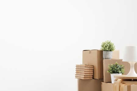 Cardboard boxes of things are stacked on the floor against a white wall. Books and table lamps and green plants in pots. The concept of moving to a new home. Copy space.の写真素材
