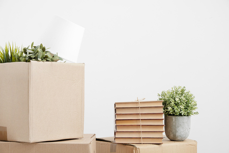 Cardboard boxes of things are stacked on the floor against a white wall. Books and table lamps and green plants in pots. The concept of moving to a new home. Copy space.の写真素材
