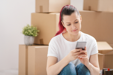 Young beautiful girl with colored hair in a white T-shirt and jeans, talking on the phone and writes messages against the background of cardboard boxes and things. The concept of transporting things on the day of moving to a new home.の写真素材