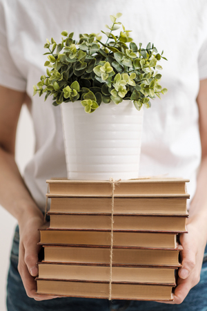 The concept of relocation and moving to a new home. Close-up, female hands hold a pile of books and a green plant in a pot. On the background of cardboard boxes and a table lamp.の写真素材