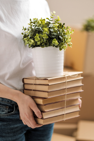 The concept of relocation and moving to a new home. Close-up, female hands hold a pile of books and a green plant in a pot. On the background of cardboard boxes and a table lamp.の写真素材