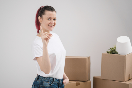 A young beautiful brunette girl in a white T-shirt holds in her hands the keys to a new home, against the background of cardboard boxes with things. The concept of moving to a new home.の写真素材