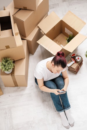 Young beautiful girl with colored hair in a white T-shirt and jeans, talking on the phone and writes messages against the background of cardboard boxes and things. The concept of transporting things on the day of moving to a new home.の写真素材