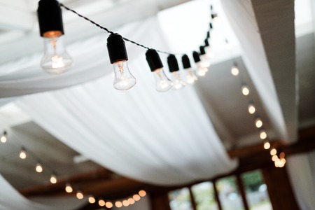 A garland of electric vintage bulbs in black cartridges is suspended from the ceiling of a light banquet hall with a white fabric awning. Concept of restaurant decor and event decoration.の写真素材
