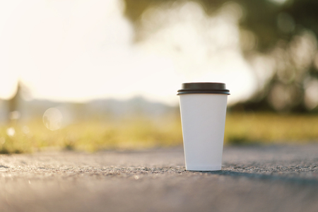 White paper cup of coffee with a lid on the ground, asphalt. Summer day, rays and flare from the sun. Warm tonality of colors. The concept of rest and relaxation in the fresh air.の写真素材