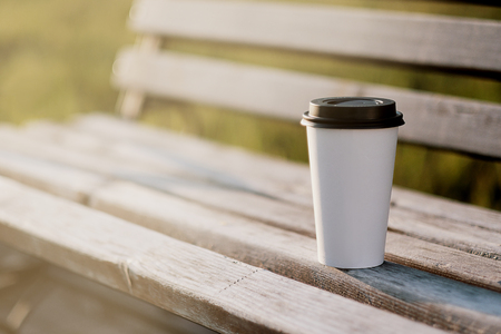 A white paper cup of coffee with a lid stands on a park bench. Summer day, rays and flare from the sun. Warm tonality of colors. The concept of rest and relaxation in the fresh air.の写真素材