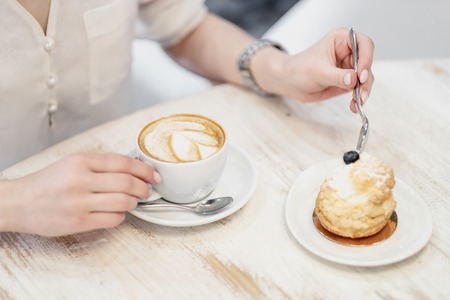 Cappuccino coffee with cup with crema in a heart wolf and cream cake dessert on a white wooden table in a cafe. Female hands with a spoon. Concept of lunch or coffee break in a restaurant.の写真素材
