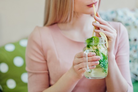 Young beautiful blonde girl in a pink dress sits at a cafe table, drinks lemonade from a glass jar and uses a smartphone. Close up lips and hands.の写真素材