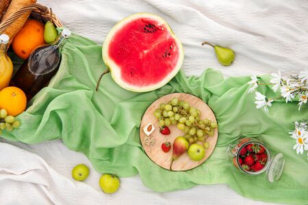Summer picnic in the meadow on the green grass. Fruit basket, juice and bottled wine, watermelon, strawberries in a glass jar and baguettes of bread. White tablecloth and a bouquet of field daisies. The concept of leisure and eating in nature.の写真素材