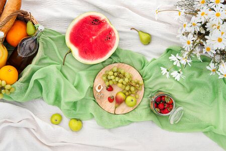 Summer picnic in the meadow on the green grass. Fruit basket, juice and bottled wine, watermelon, strawberries in a glass jar and baguettes of bread. White tablecloth and a bouquet of field daisies. The concept of leisure and eating in nature.の写真素材