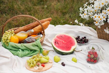 Summer picnic in the meadow on the green grass. Fruit basket, juice and bottled wine, watermelon and bread baguettes. White tablecloth and a bouquet of field daisies. The concept of recreation and food in nature.の写真素材