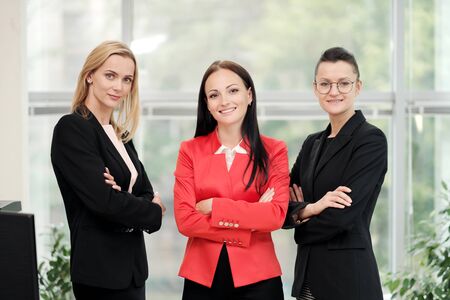 Three young attractive women in business suits posing against the backdrop of a light office. Head and subordinates. Working team of professionals and colleagues. Feminism and feminine power.の写真素材