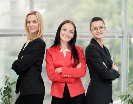 Three young attractive women in business suits posing against the backdrop of a light office. Head and subordinates. Working team of professionals and colleagues. Feminism and feminine power.の写真素材