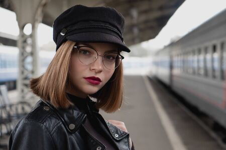Attractive young woman millenial in black clothes and a hat and glasses at the railway station next to the train. Serious look, waiting for the train. Big clock on the background.の写真素材