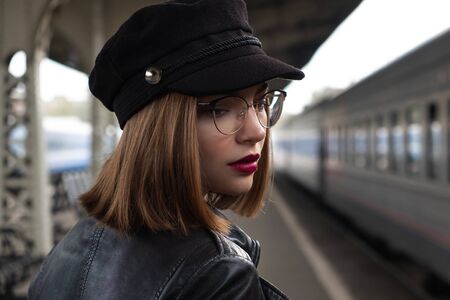 Attractive young woman millenial in black clothes and a hat and glasses at the railway station next to the train. Serious look, waiting for the train. Big clock on the background.の写真素材