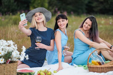 Three young women, in blue dresses and hats sit on plaid and take pictures on smartphone. Outdoor picnic on grass. Delicious food in picnic basket and wine. Watermelon, grapes and bouquet of daisies.の写真素材