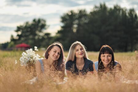 Three young women in blue dresses, and hats lie on plaid and drink wine. Outdoor picnic on grass on beach. Delicious food in picnic basket and wine. Watermelon, grapes and bouquet of daisies.の写真素材