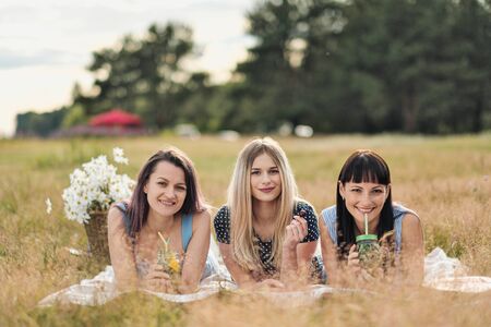 Three young women in blue dresses, and hats lie on plaid and drink wine. Outdoor picnic on grass on beach. Delicious food in picnic basket and wine. Watermelon, grapes and bouquet of daisies.の写真素材