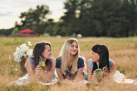 Three young women in blue dresses, and hats lie on plaid and drink wine. Outdoor picnic on grass on beach. Delicious food in picnic basket and wine. Watermelon, grapes and bouquet of daisies.の写真素材