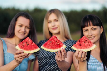 Three young attractive women friends in blue dresses at sunset are eating watermelon and smiling. Outdoor picnic.の写真素材
