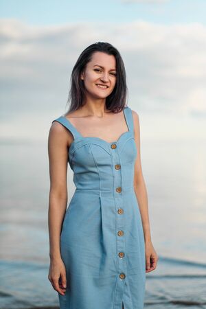 A young attractive dyed hair woman, in a blue dress, walks along the seashore. Portrait of a smiling woman on vacation.の写真素材