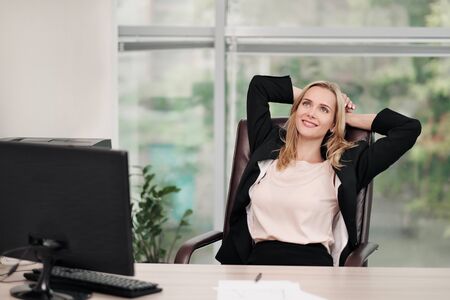 A young attractive woman in a business suit sits at a desk. Stretches and relaxes on the chair. Feet are on the table. Rest from work. Fatigue, a break in work.の写真素材