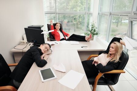 Three beautiful young girls in business suits are sitting at the office desk. Tired of work. Throw paper airplanes, and laugh. Break and rest in the workplace.の写真素材