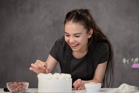 An attractive young female brunette confectioner decorates a white cake with small red flowers of...