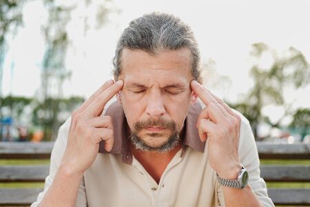 A tired and distressed aged man sits on a park bench. Leans on his hands, rubs his temples....