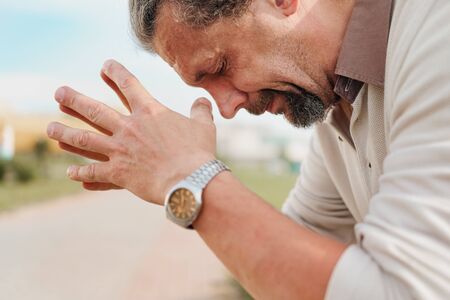 A tired and distressed aged man sits on a park bench. Leans on his hands, rubs his temples. Headache.の写真素材