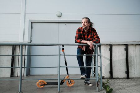 An elderly man with long hair and a beard rides a scooter through the city streets. Check shirt and...