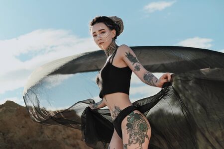 Beautiful sexy woman in black underwear. The body is covered with many tattoos. On the head dreadlocks. Posing against a sandy career and black fabric.の写真素材
