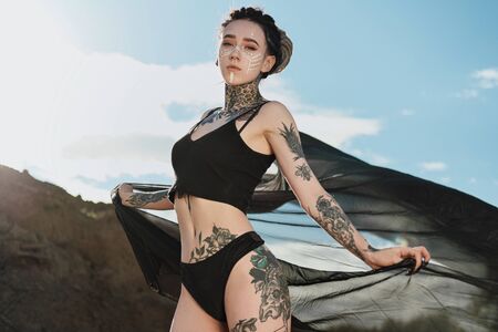 Beautiful sexy woman in black underwear. The body is covered with many tattoos. On the head dreadlocks. Posing against a sandy career and black fabric.の写真素材