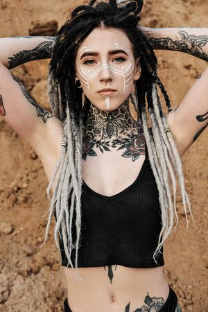 Beautiful sexy woman in black underwear. The body is covered with many tattoos. On the head dreadlocks. Posing against a sandy career and black fabric.の写真素材