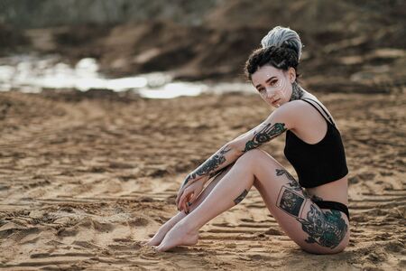 Beautiful sexy woman in black underwear. The body is covered with many tattoos. On the head dreadlocks. Posing against a sandy career and black fabric.の写真素材