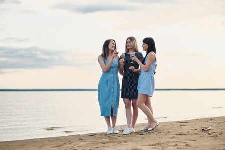 Three young attractive women, in blue dresses are walking along the seashore. Girlfriends communicate, laugh and drink wine. Outdoor recreation.の写真素材