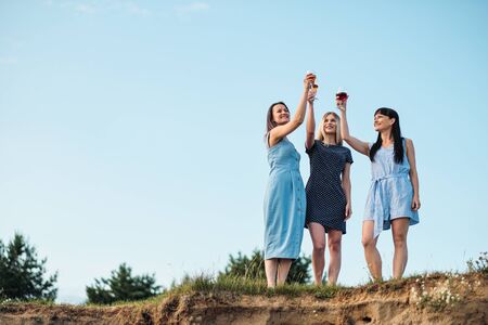 Three young attractive women, in blue dresses are walking along the seashore. Girlfriends communicate, laugh and drink wine. Outdoor recreation.の写真素材